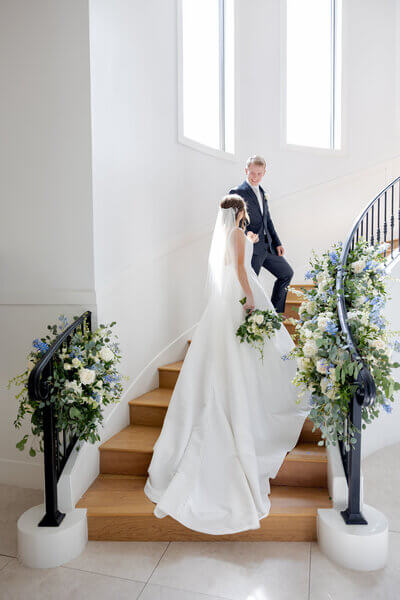 Bright photo of groom holding the brides hand and leading bride up spiral floral covered stairwell while smiling