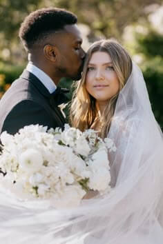 Photo of groom kissing his bride on the forehead while her veil and all white bouquet are shown in the lower half of the frame