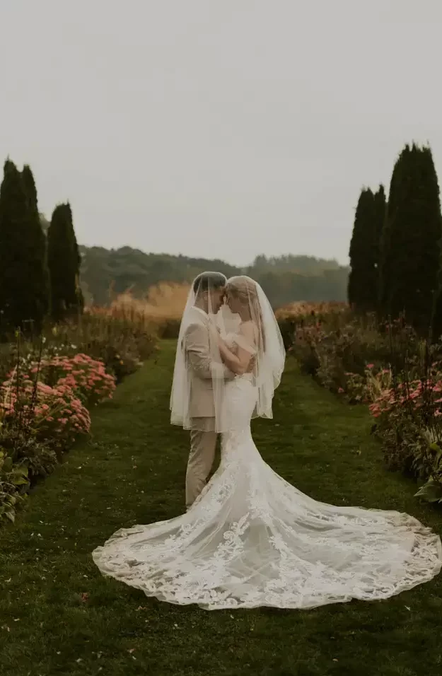 Vertical photo of bride and groom pressing their foreheads together under the brides veil in the middle of a garden pathway surrounded with shrubs and flowers