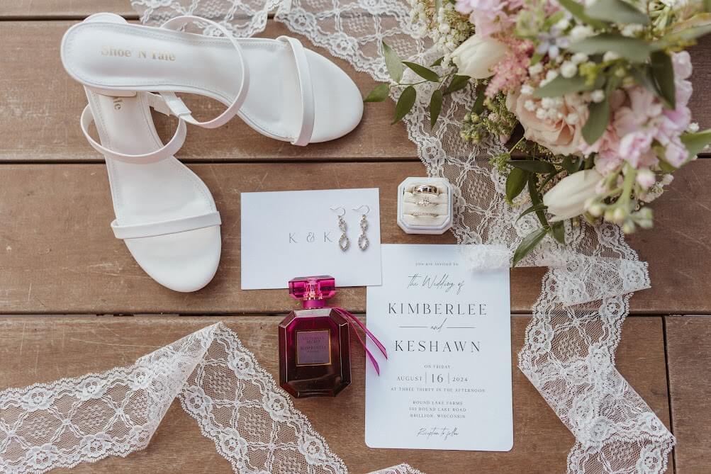 Overhead photo of brides heels, perfume, earring, rings in their box, wedding program and bouquet all surrounded by lace