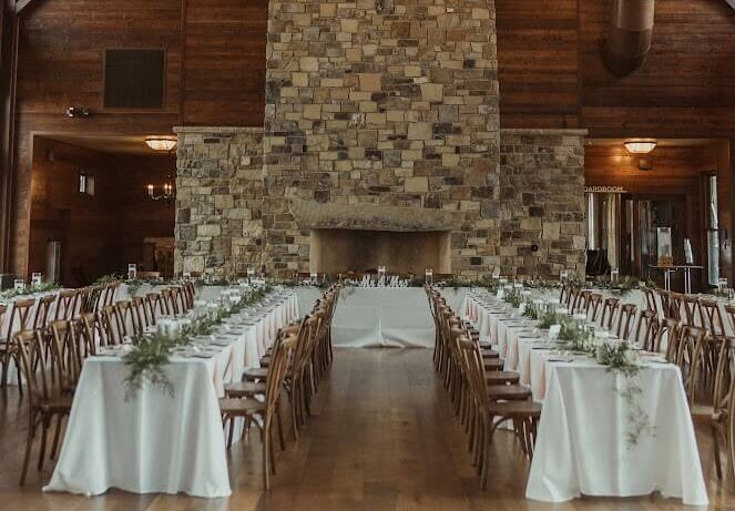 Wide photo of large warm cabin dining room with tables and chairs laid out below the large stone fireplace